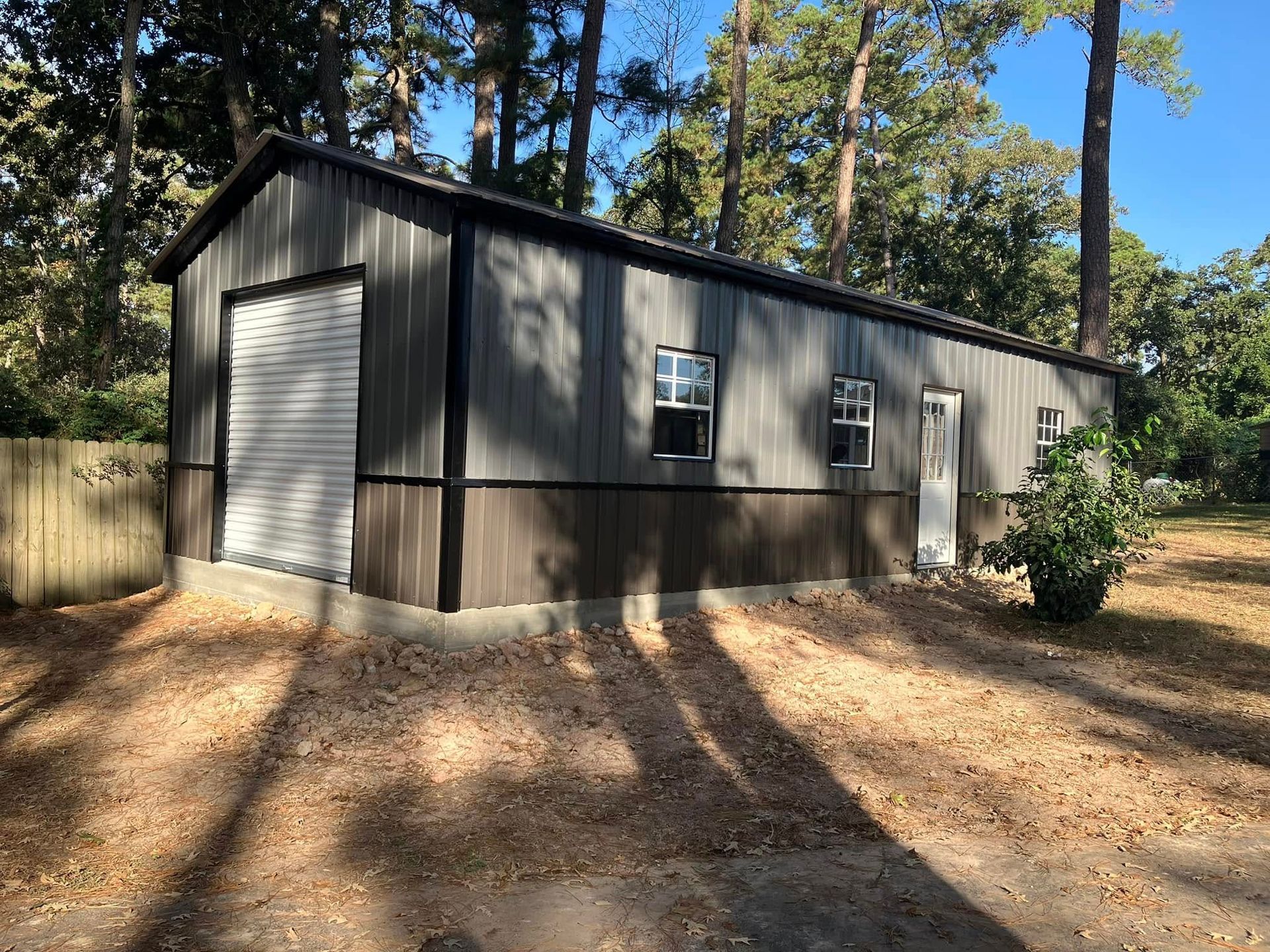 Metal garage with a roll-up door, windows, and a side door; surrounded by trees and a yard.