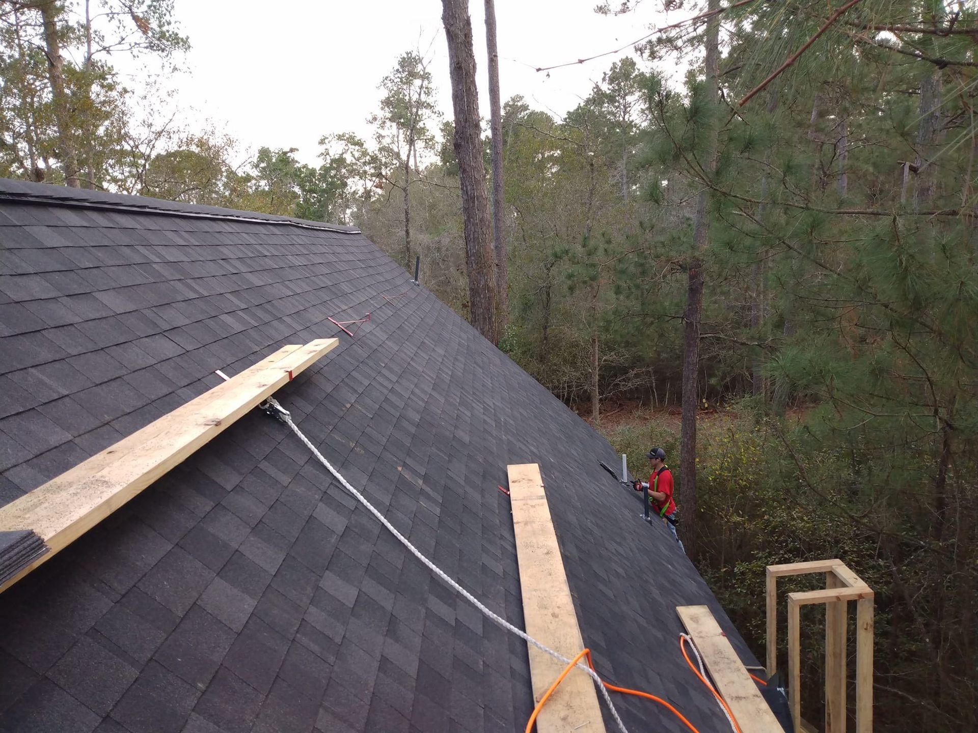 Roofer working on a dark shingled roof in a wooded area. Wood planks and safety lines are present.