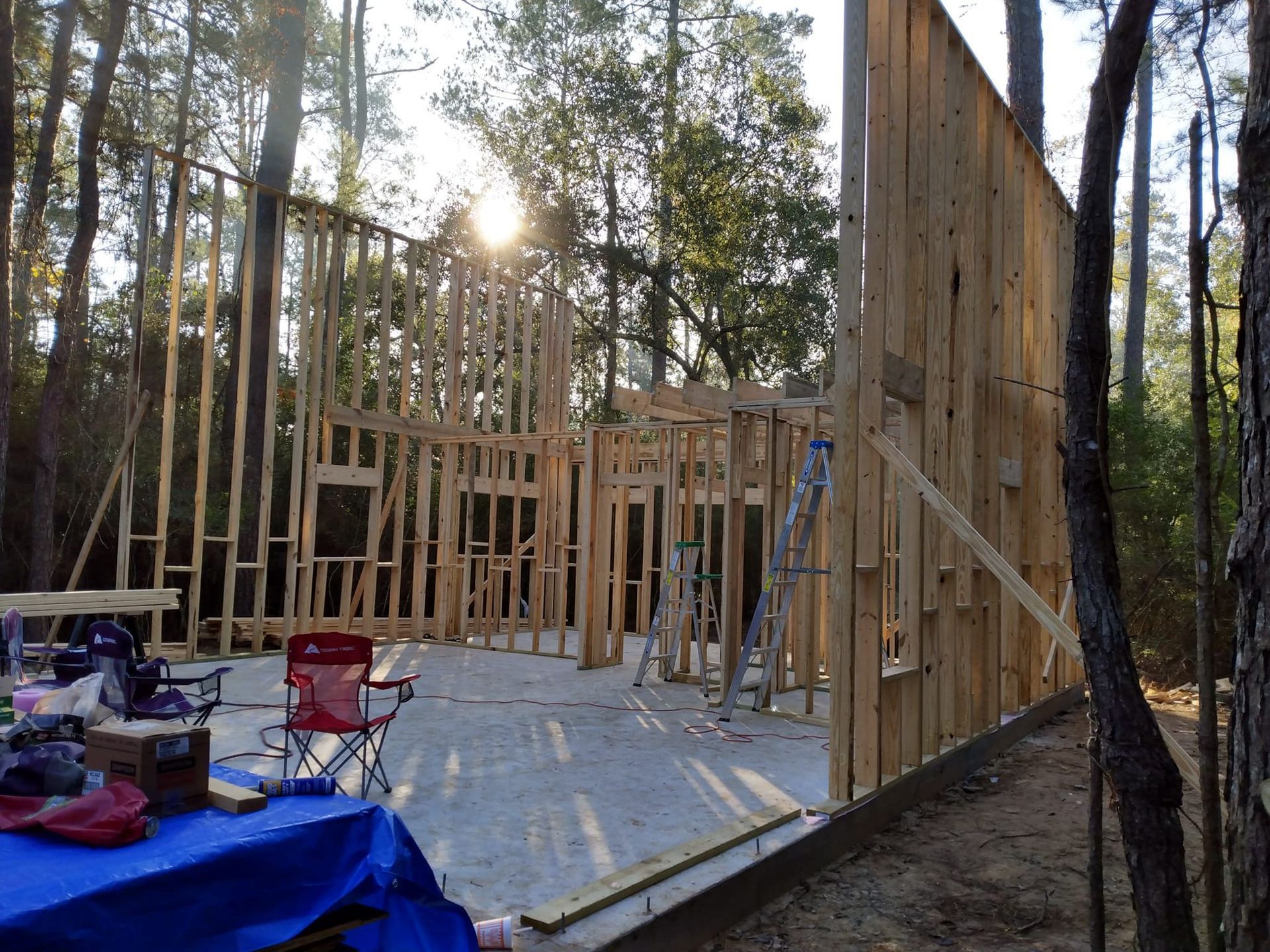 Framing of a wooden house under construction in a wooded area; sunlight shines through the incomplete frame.