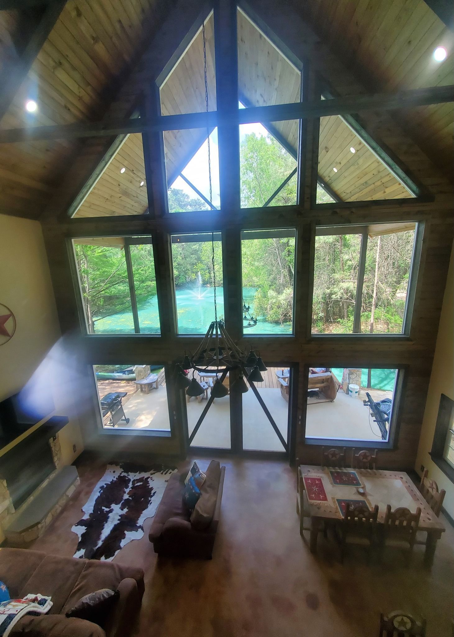 High-ceilinged cabin interior with large windows overlooking trees. Living area with fireplace, seating, and dining table.