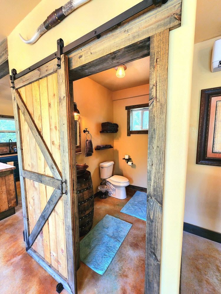 Barn door opens to a rustic bathroom with barrel sink, toilet, and blue rugs.