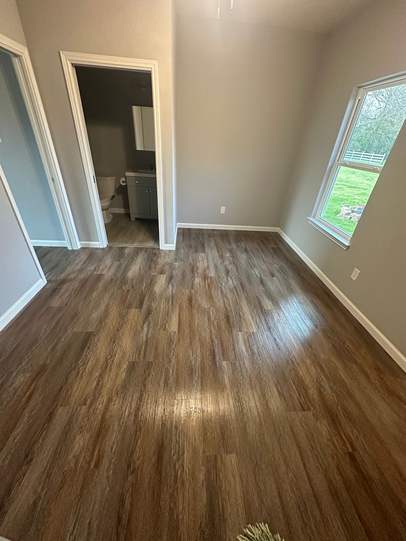 Empty room with wood-look floor, gray walls, and an open doorway leading to a bathroom. A window provides natural light.