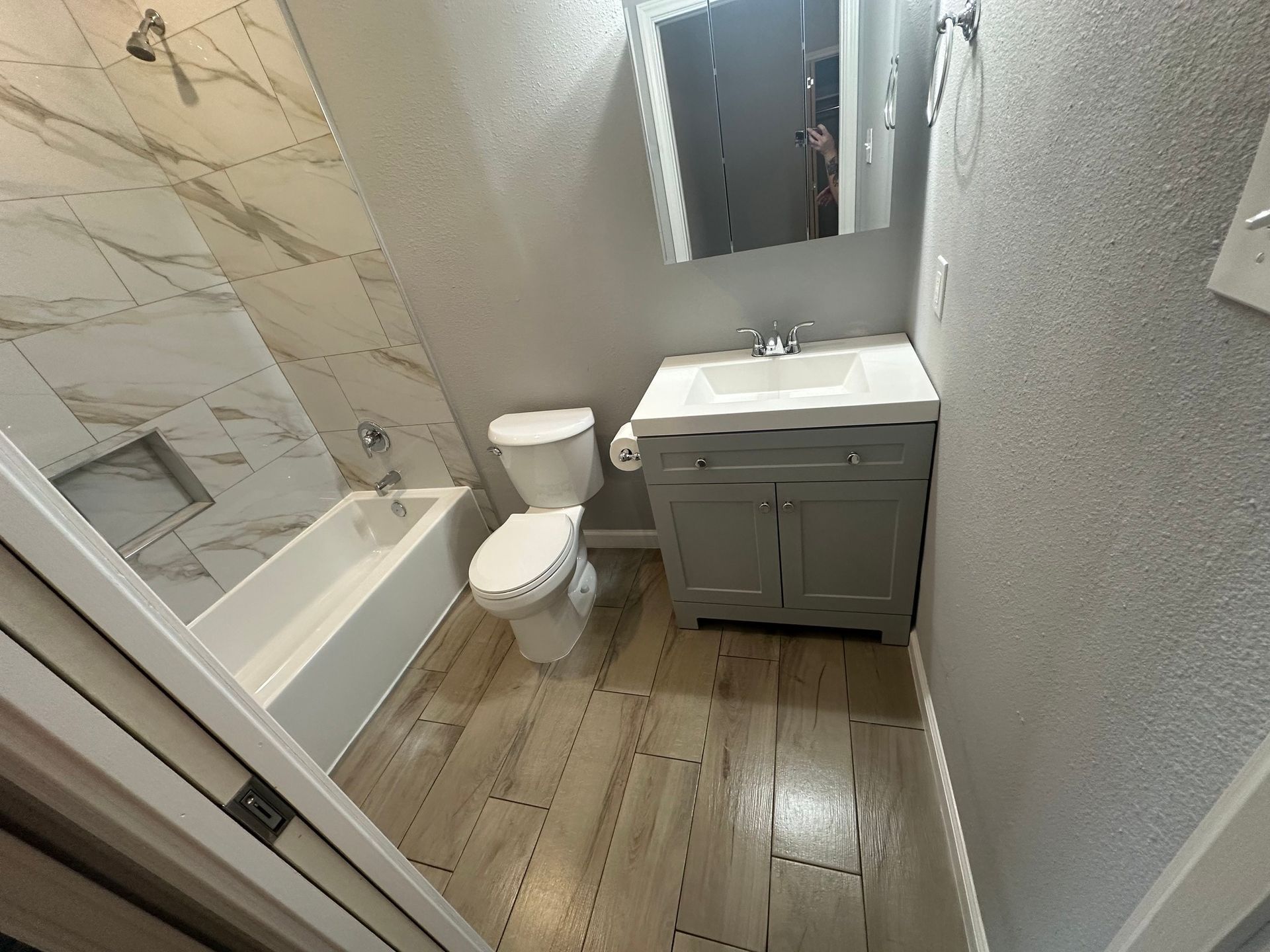 A bathroom with light wooden floor tiles, grey vanity, white toilet, and marble-patterned shower tiles.