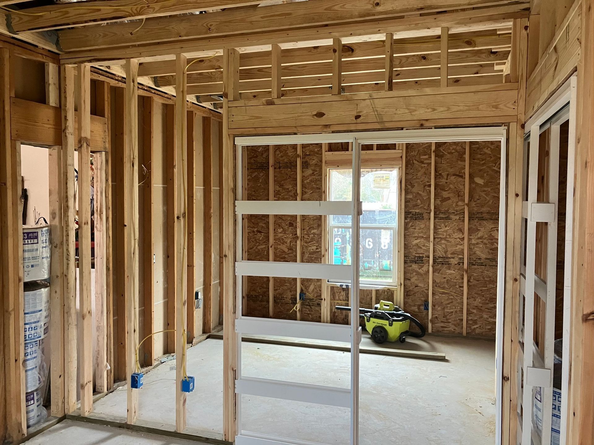 Interior view of a room under construction, showing wooden wall frames, a doorway, and a window with a view.