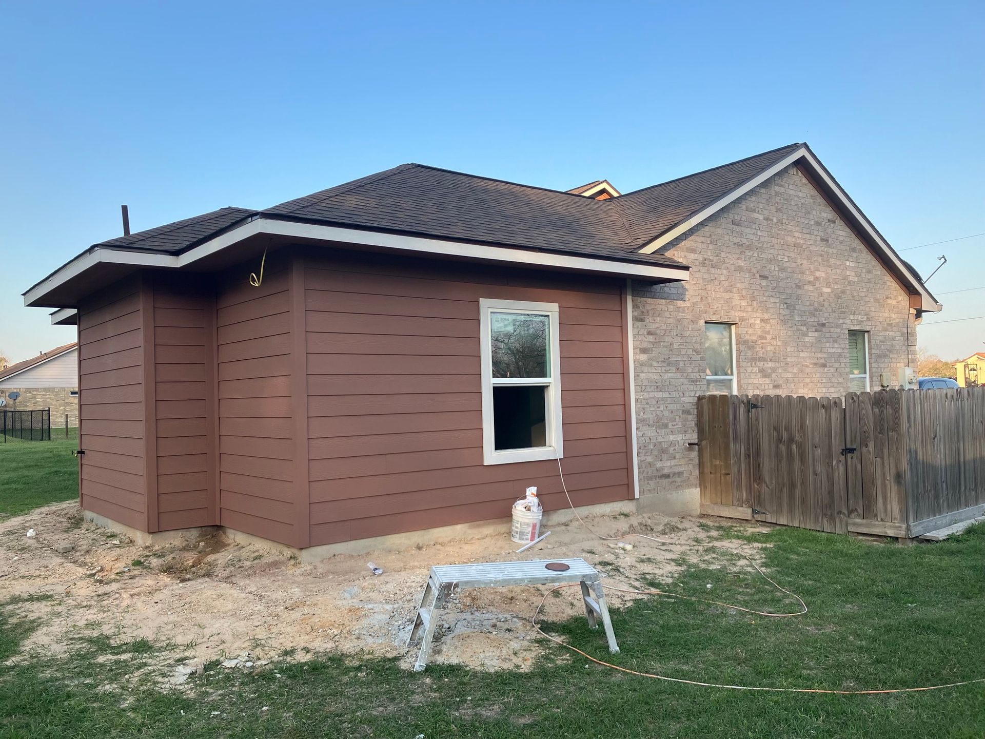 Brown-sided addition to a brick house with a window, next to a wooden fence and grass, with a blue sky.