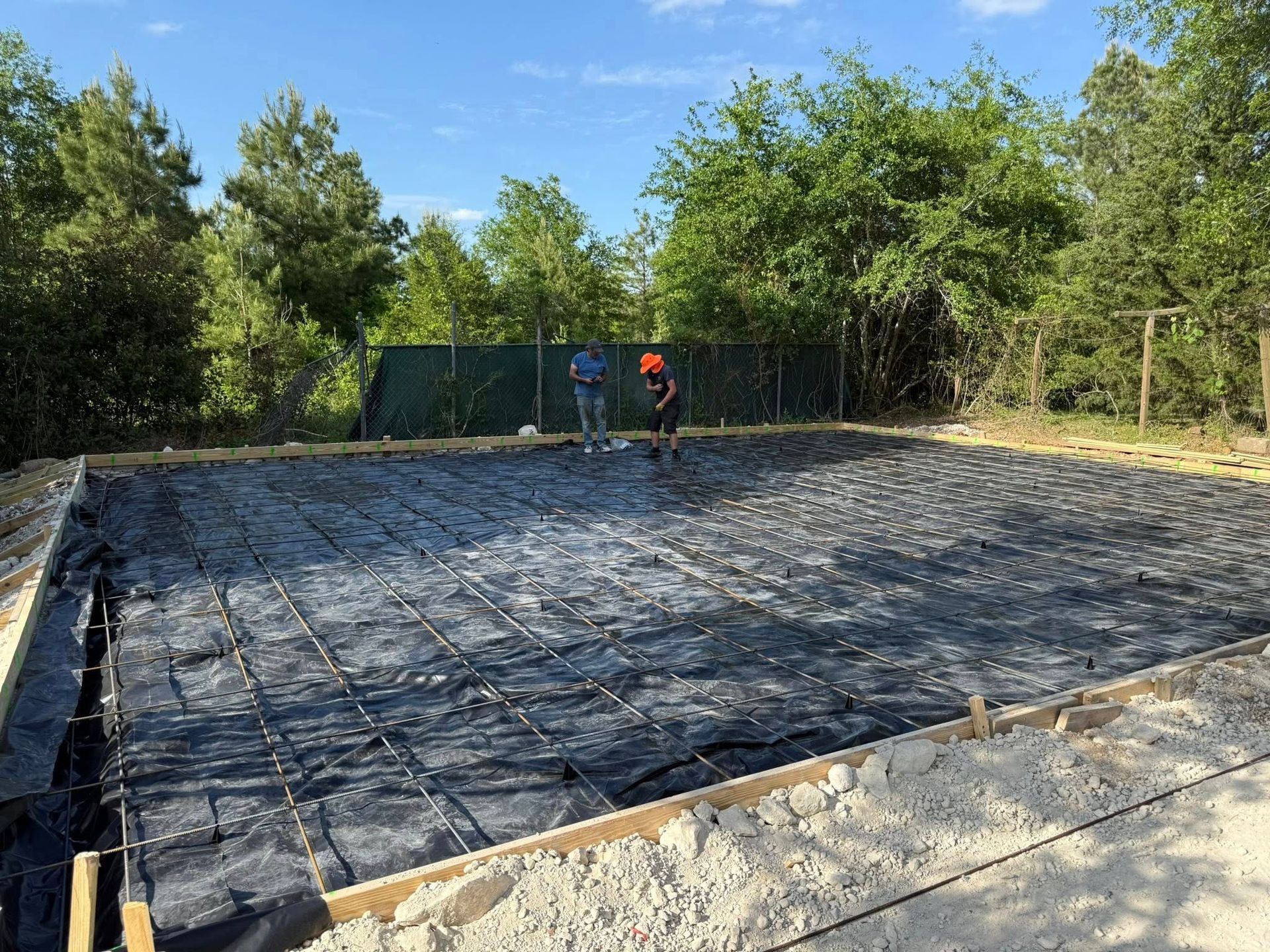 Construction site: concrete foundation with rebar grid and two workers, green trees in background.