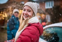 A young woman with braces on her teeth is standing in front of a car in the snow.
