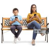 A woman and a boy are sitting on a bench texting on their phones