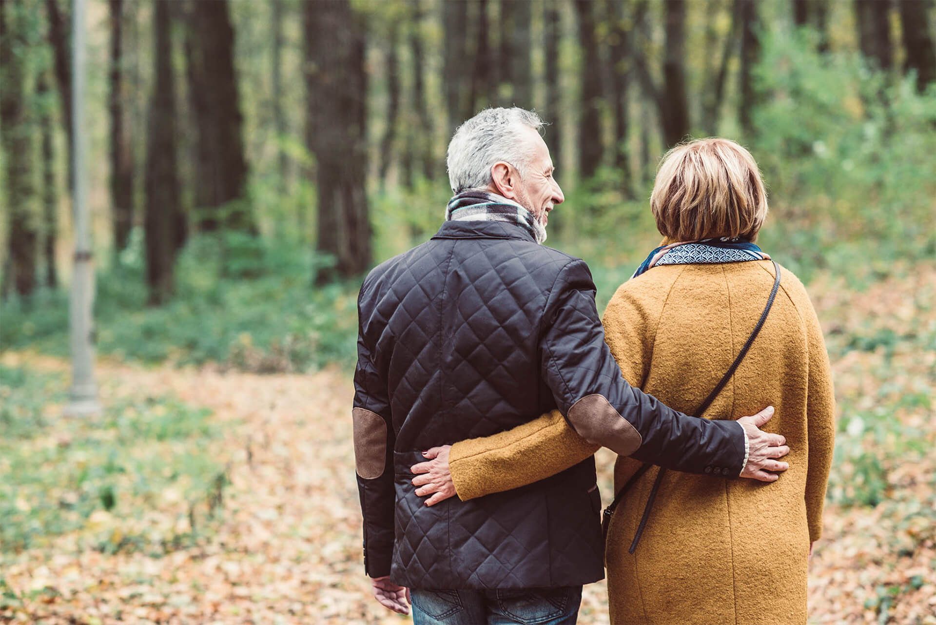 Couple walking in a forest, man's arm around woman's shoulder. Autumn leaves, trees in background.