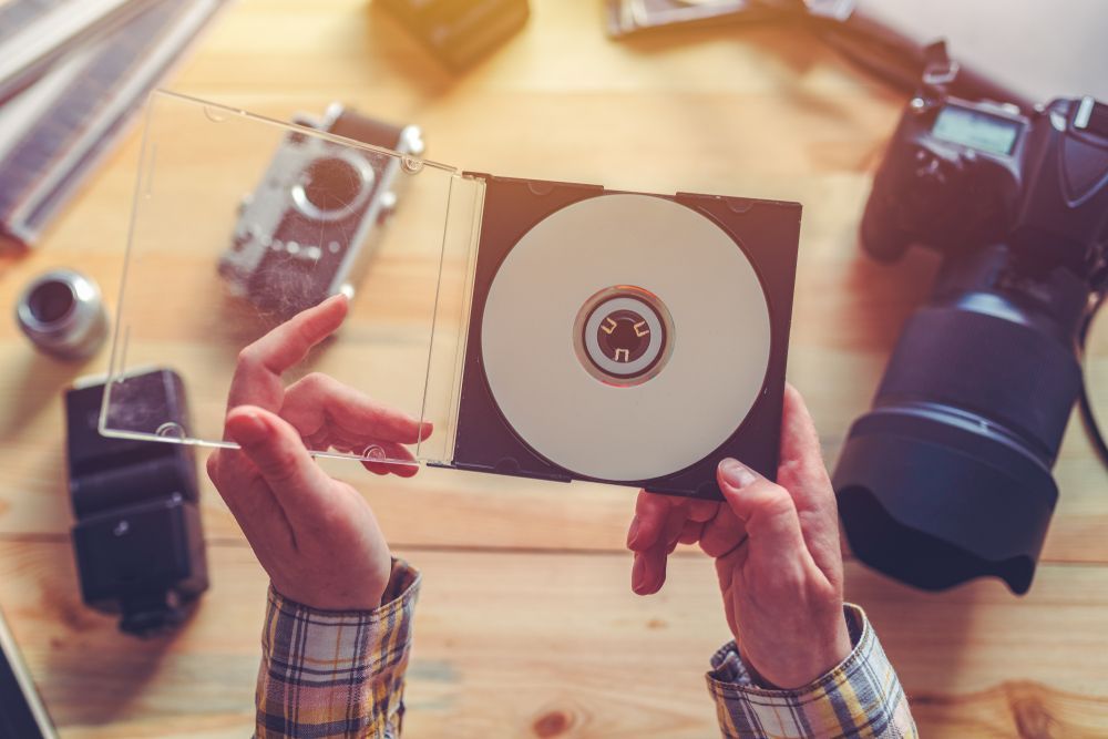 Hands holding a CD in its case, surrounded by cameras and photography equipment on a wooden table.