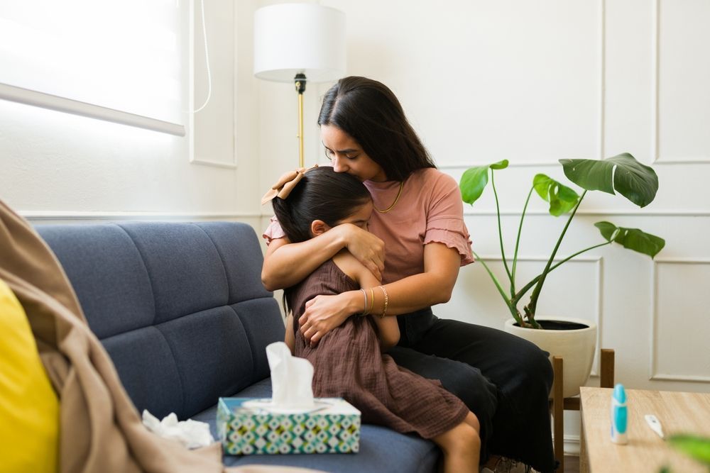 Woman comforting child on a blue couch, embracing and touching the child's head, tissues nearby.