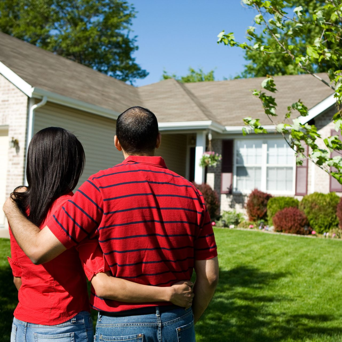 Couple embracing, looking at a house with a green lawn and trees on a sunny day.