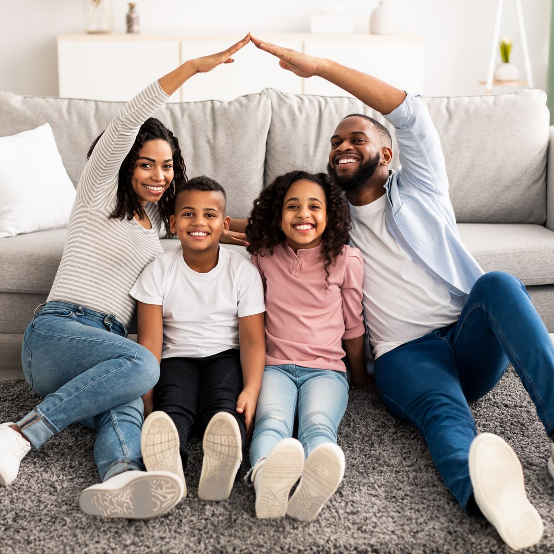 Family of four smiling under a roof shape made with their arms, sitting on a carpet in front of a couch.