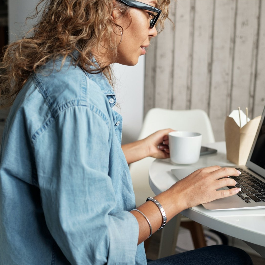 Woman in blue shirt working on laptop, holding mug, indoors.