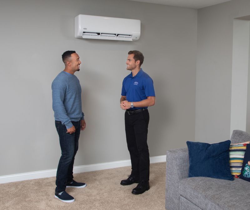Man and HVAC technician discuss an air conditioner mounted on a gray wall in a living room.