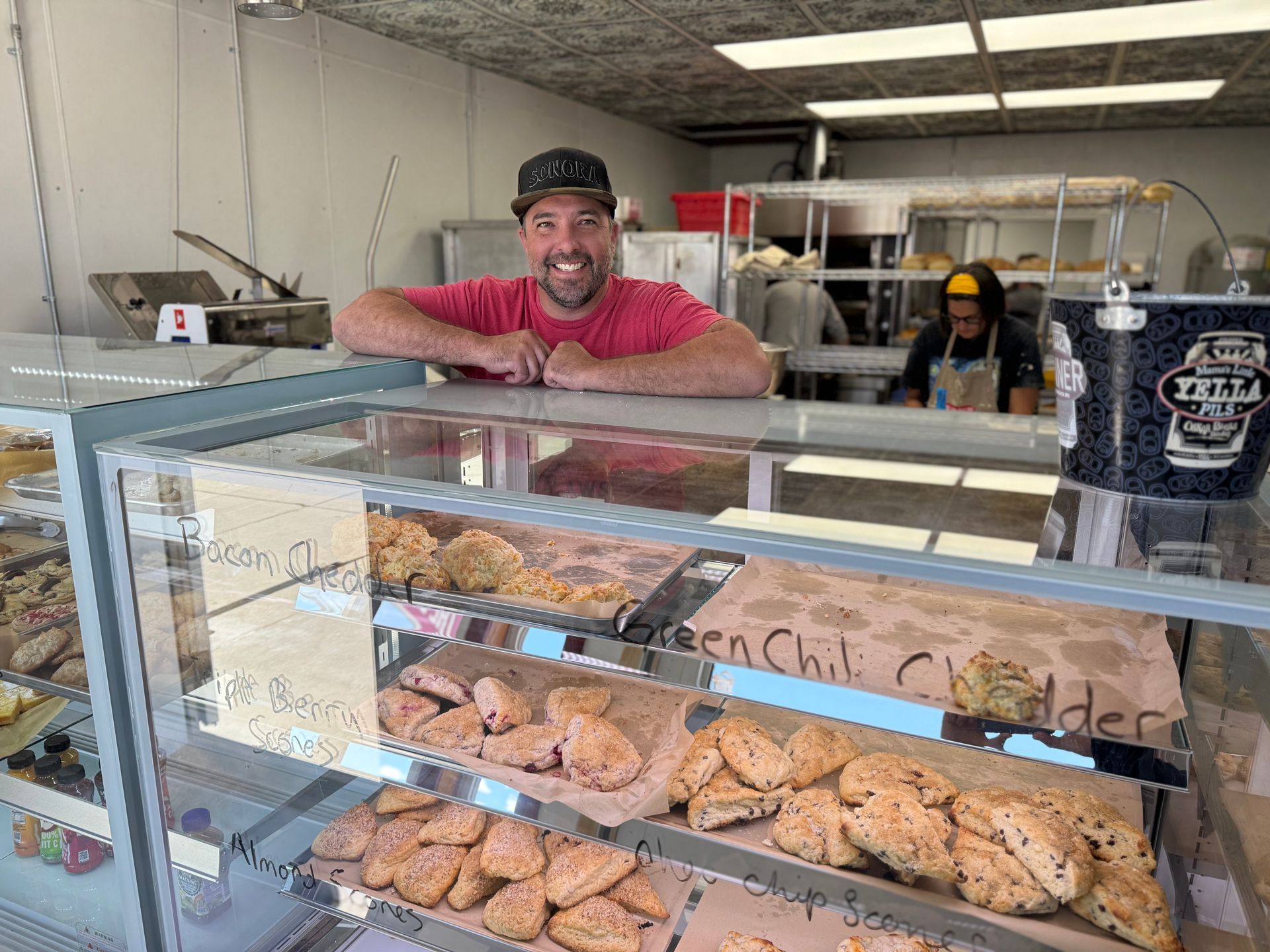A man is leaning on a glass display case in a bakery.