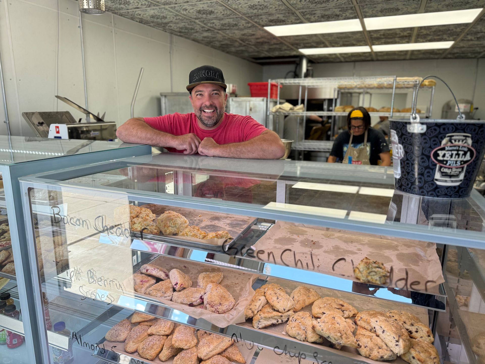A man is leaning on a glass display case in a bakery.