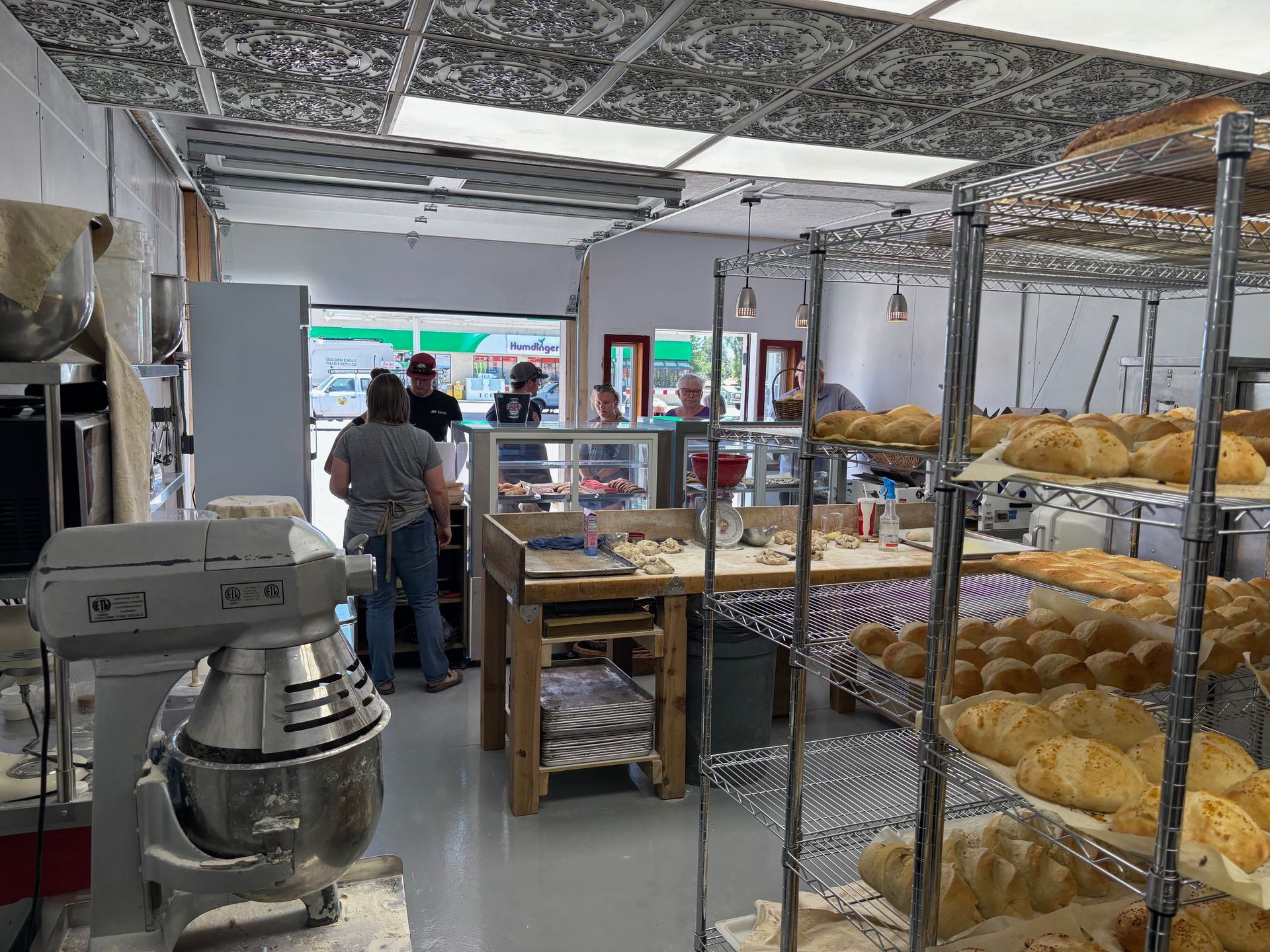 Bakery interior with metal shelves of bread, a mixer, people at the counter, and a doorway to the outside.