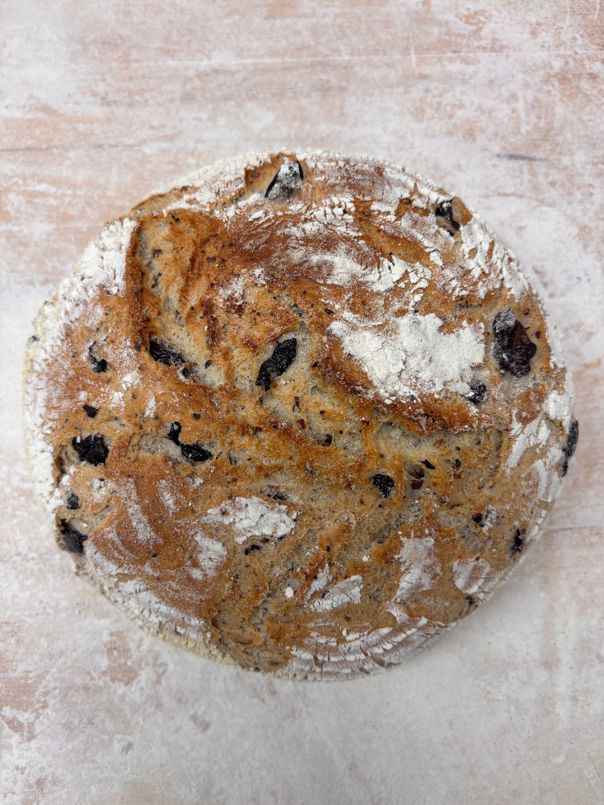 Round loaf of olive bread on a light wood surface, dusted with flour.
