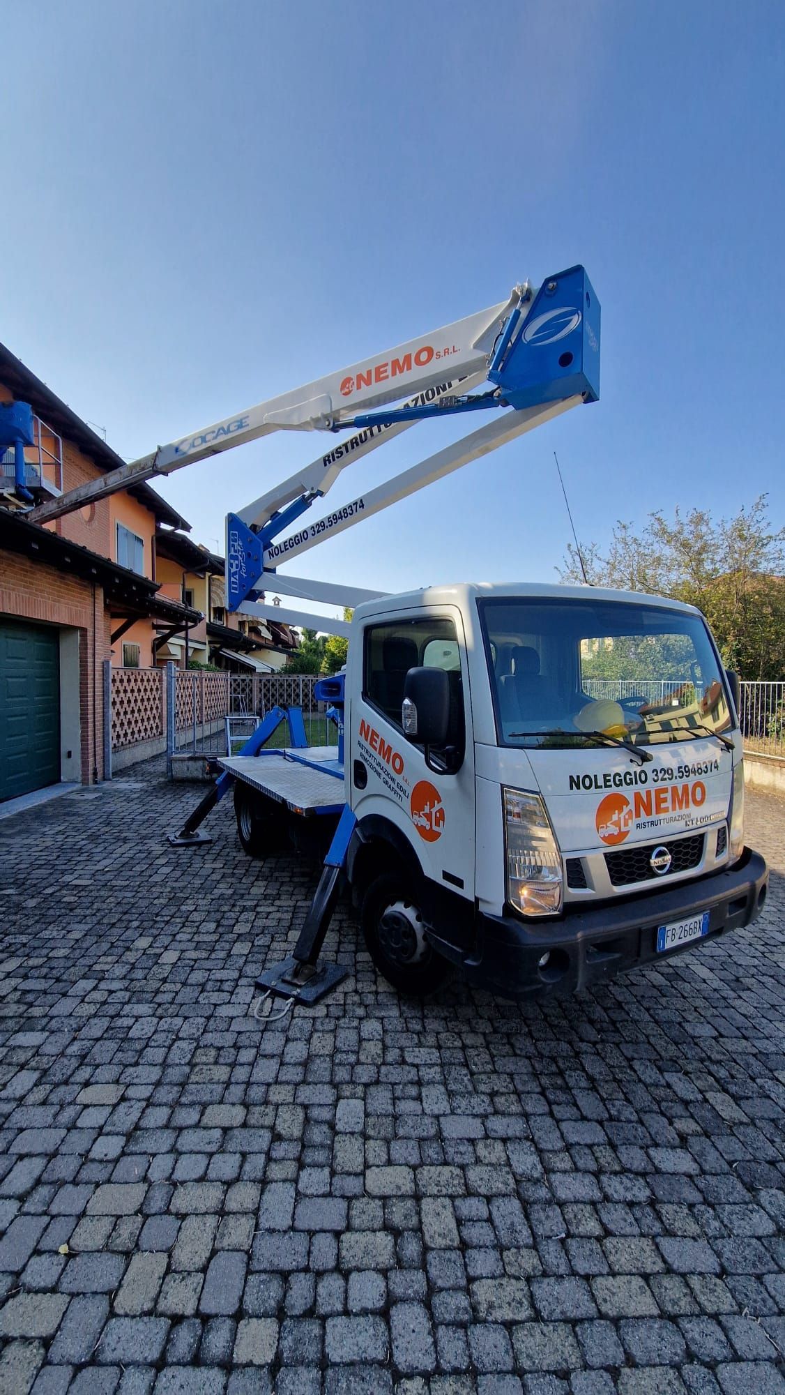Camion bianco con piattaforma aerea estesa; parcheggiato sul selciato. La piattaforma è blu e bianca, vicino a una casa.