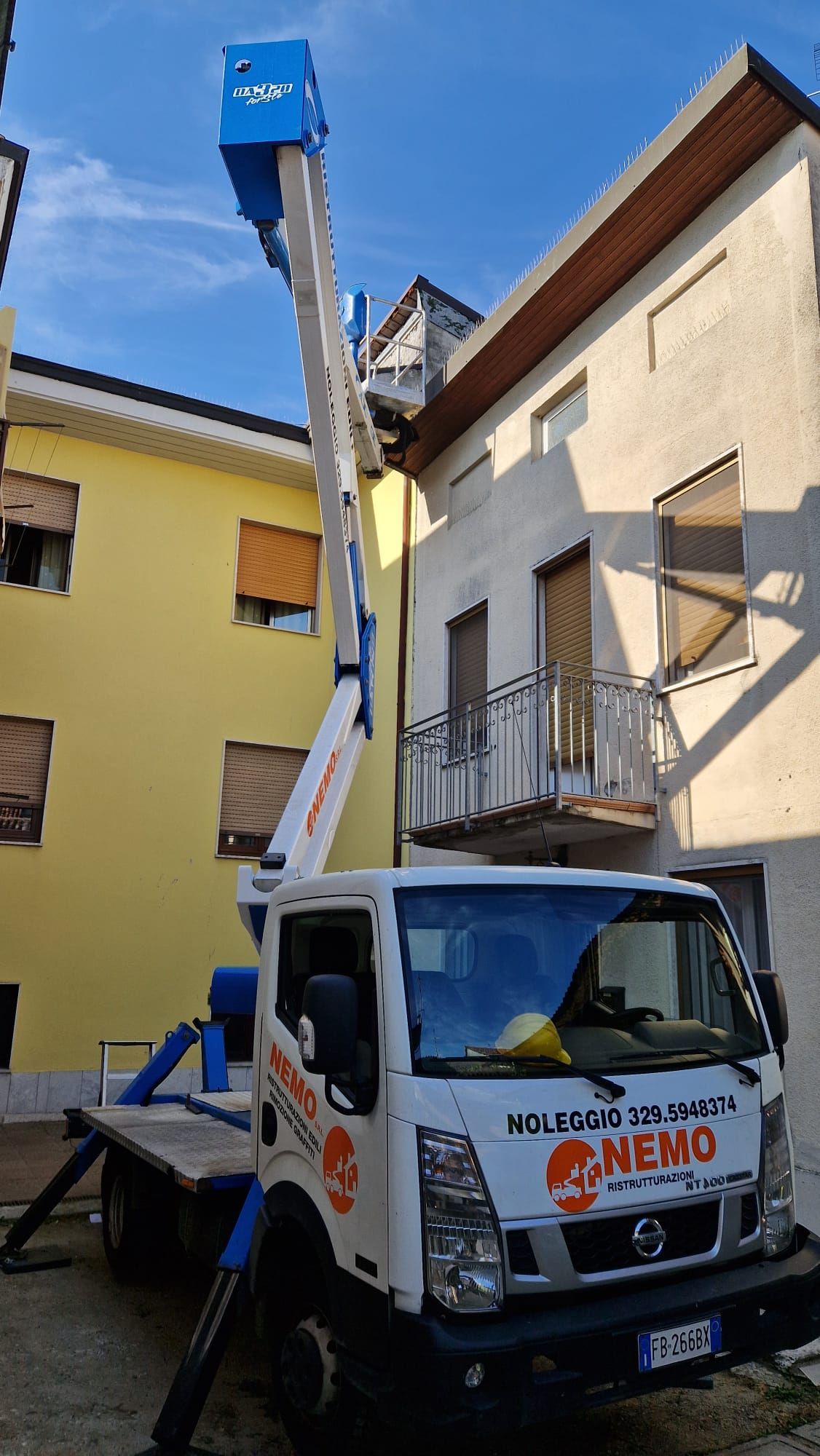 Una piattaforma aerea montata su un camion in uno stretto vicolo tra edifici gialli e bianchi. Il braccio della piattaforma è proteso verso un tetto.