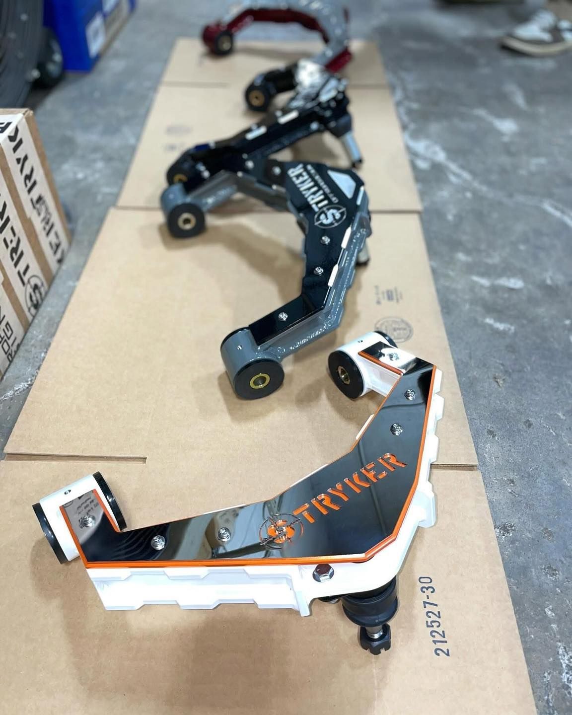 Five automotive suspension control arms, various colors, lined up on cardboard.