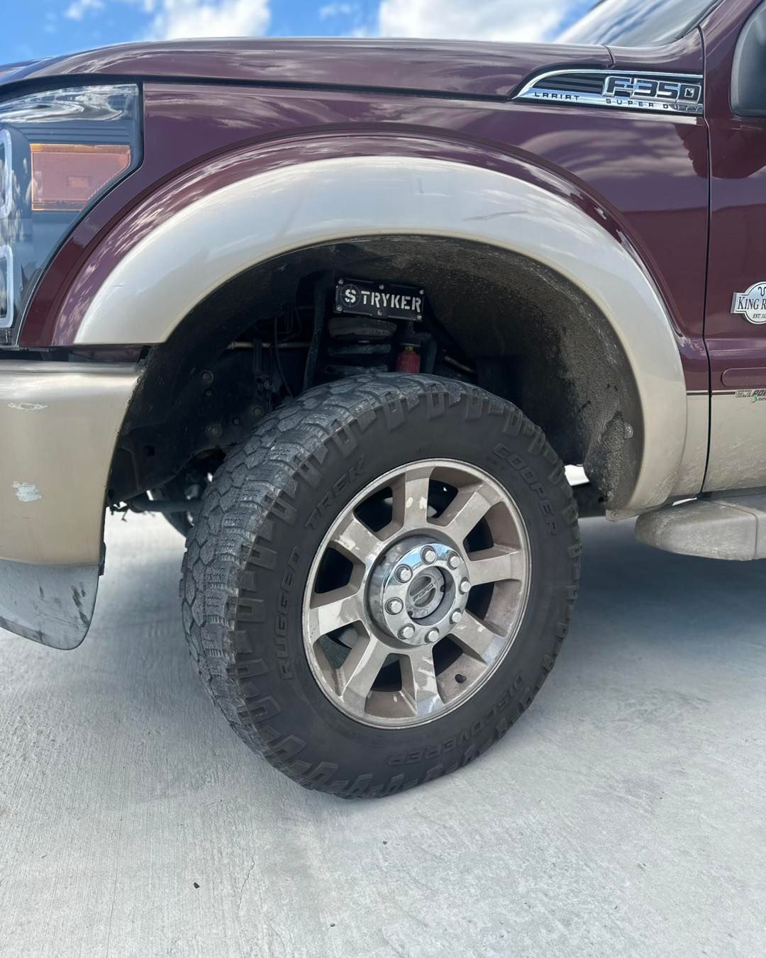A close-up of a burgundy and tan Ford truck's front wheel. Tire is dark with tan rim and a light brown wheel well.