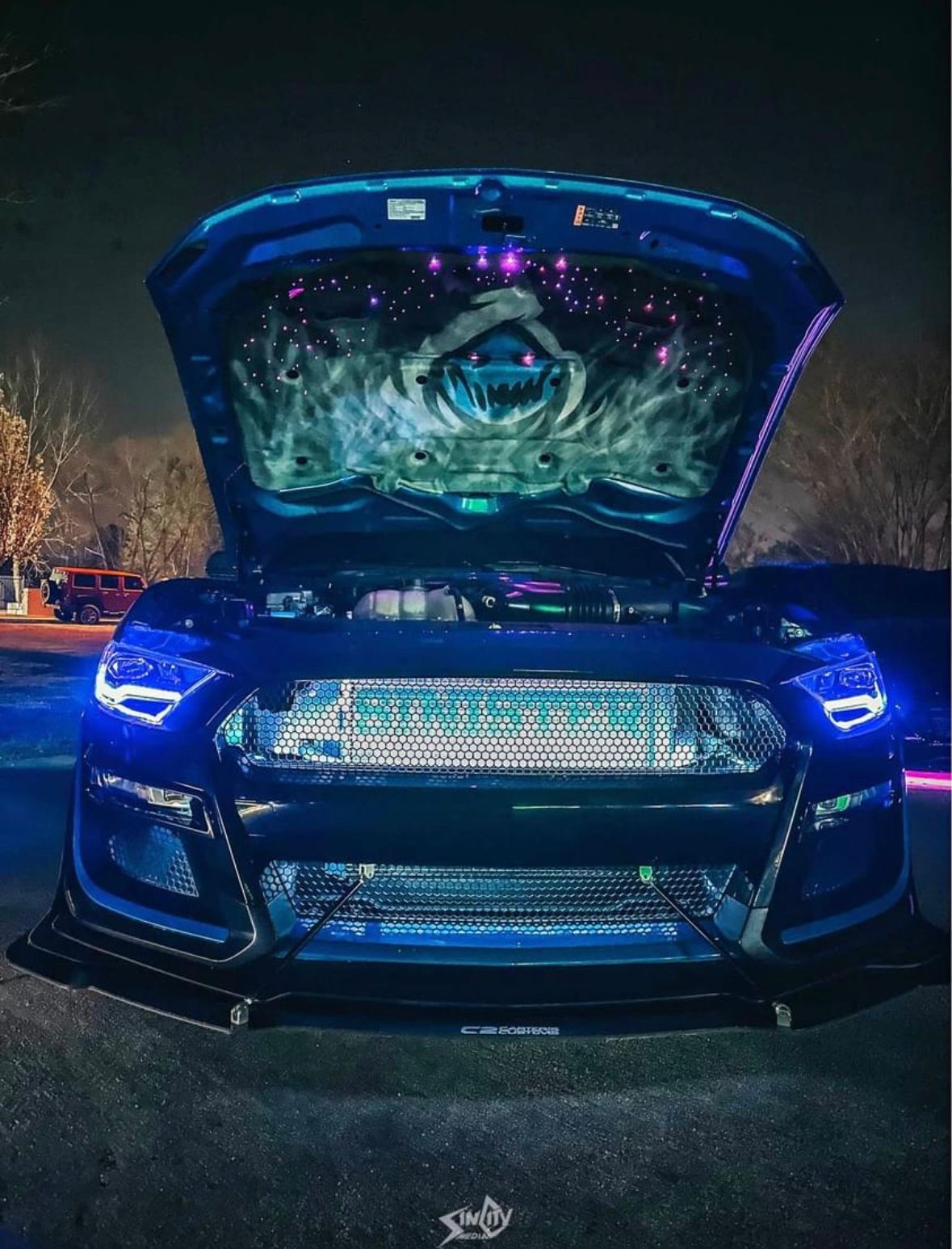 Blue customized Mustang with hood open, lit up with neon blue lights. Painted design on hood.