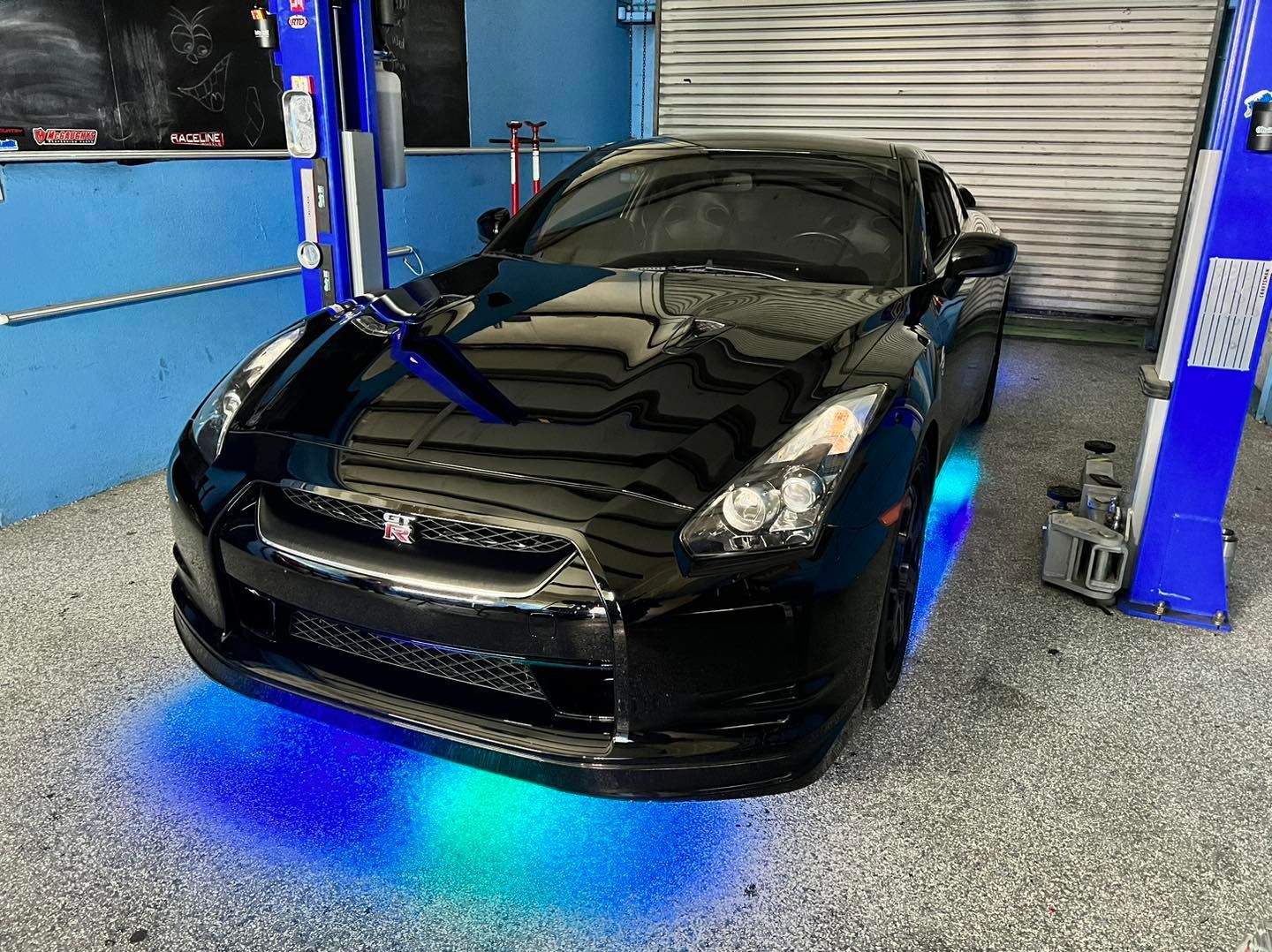 Black Nissan GT-R in garage with blue underglow, between car lifts.
