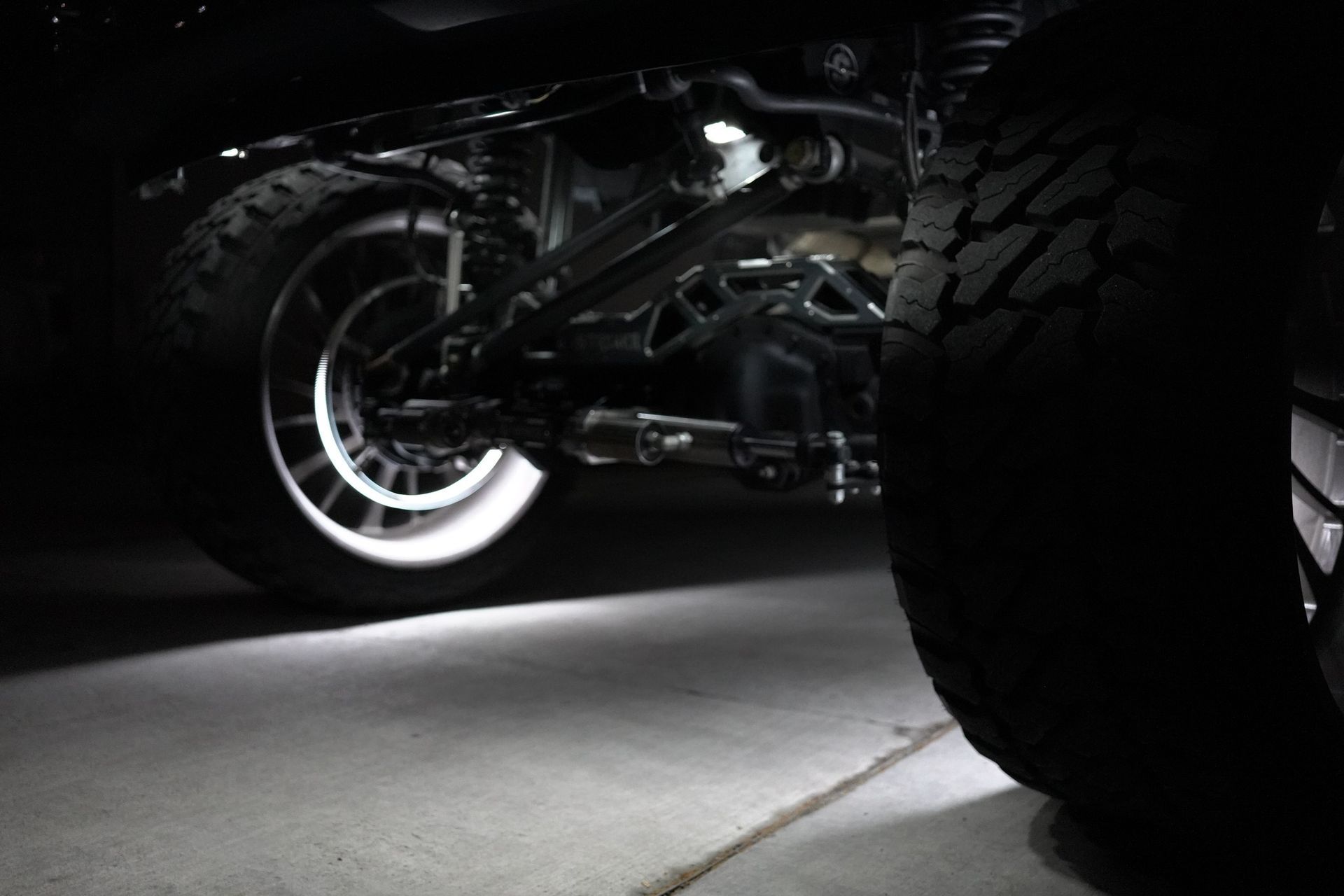 Dark, undercarriage of a vehicle with visible tires and illuminated underglow lights on concrete.
