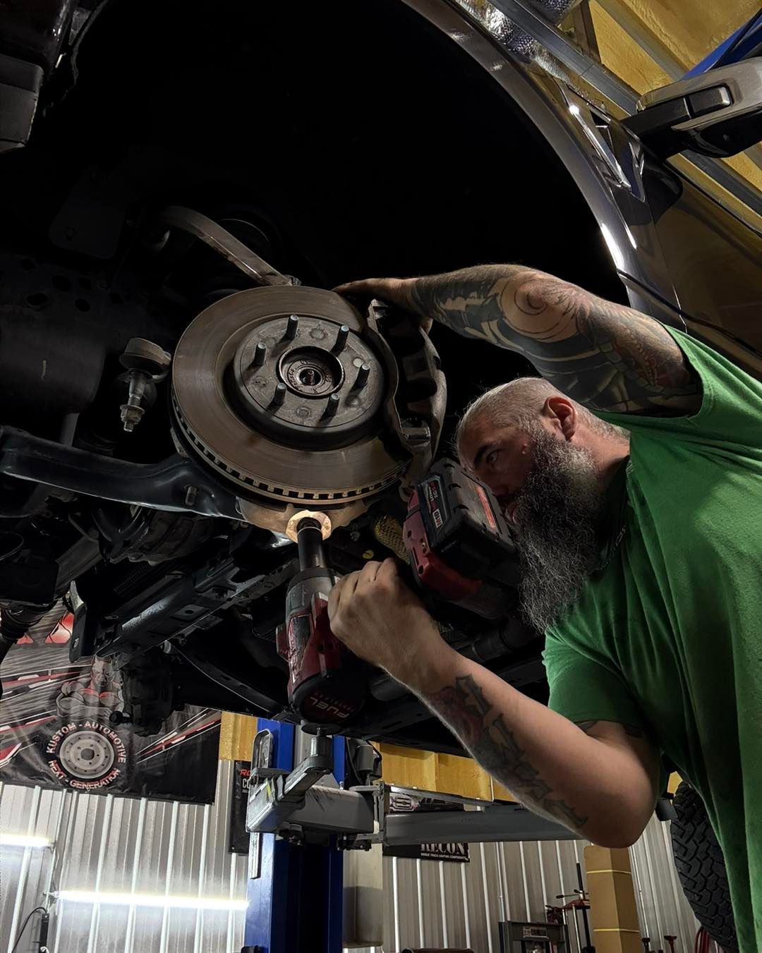 Mechanic working on car brakes with a power drill.  Garage setting, bearded man, green shirt.