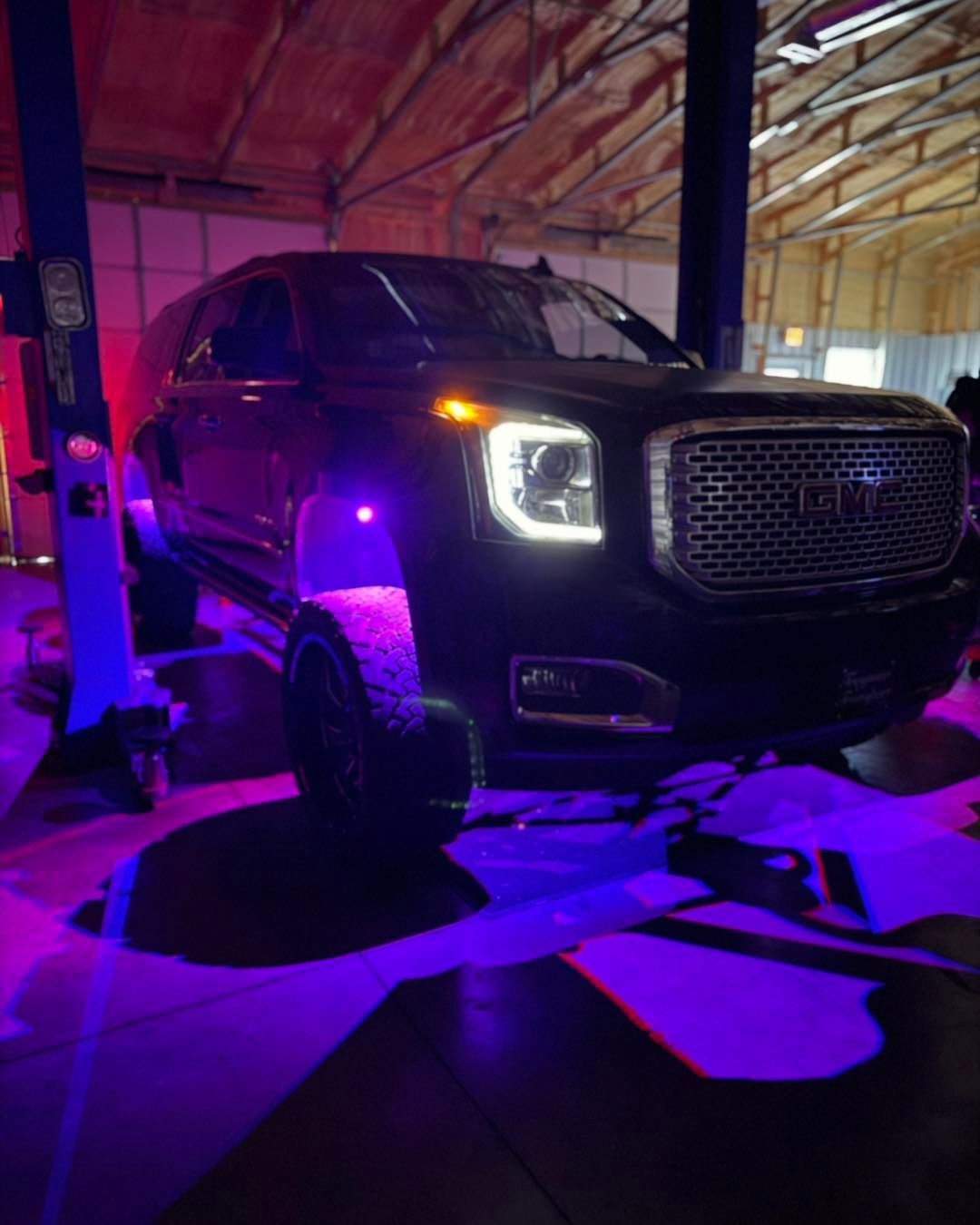 Black GMC Yukon on a car lift in a garage, illuminated by purple and red lights.