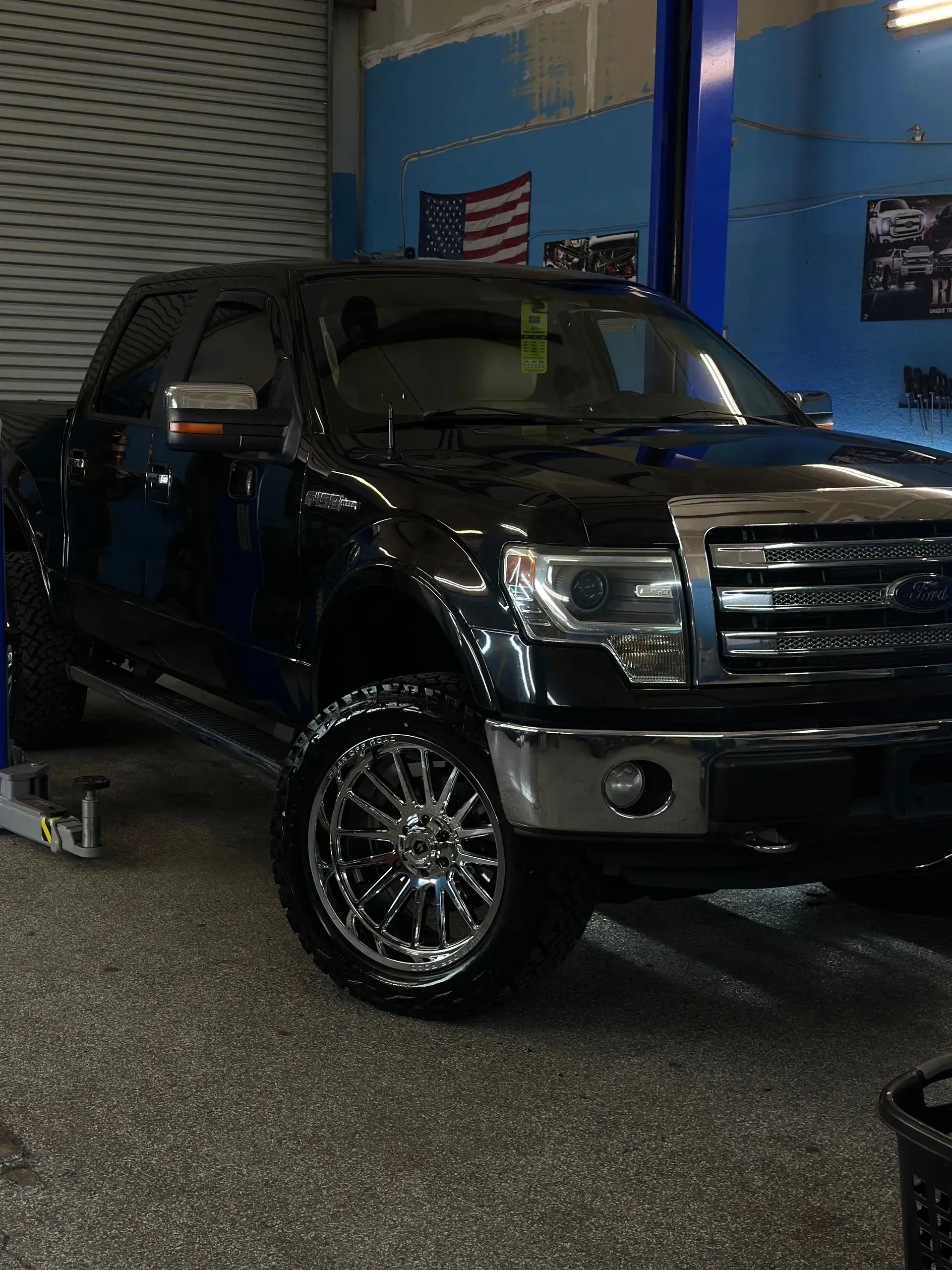 Black Ford F-150 truck with chrome rims in an auto repair shop, with an American flag in background.