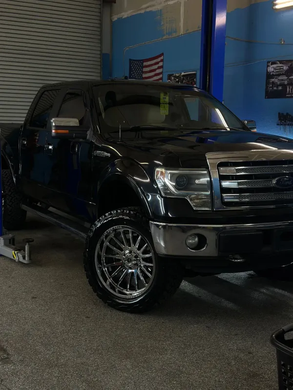 Black Ford F-150 truck with chrome rims in an auto repair shop, with an American flag in background.