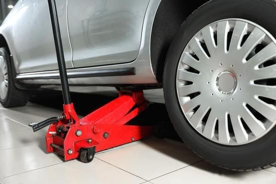 Red car jack lifting a silver car's tire, indoors on tiled floor.