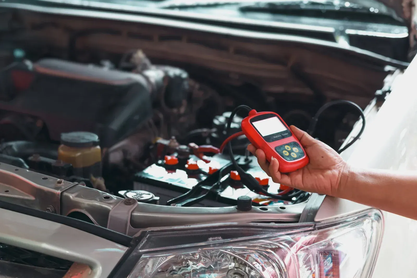 A person tests a car battery with a red and black digital reader in an open engine bay.