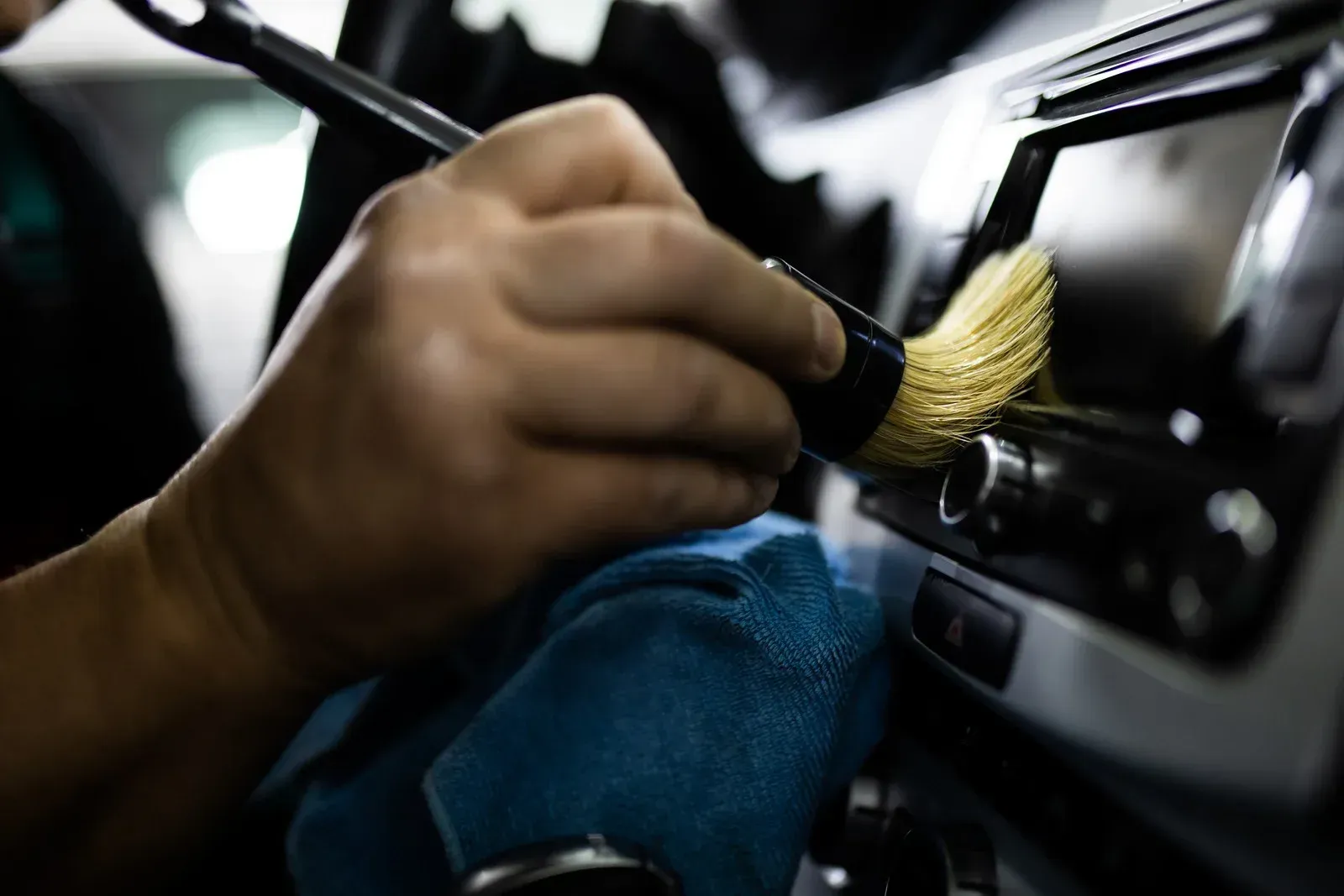 Hand using brush to dust car dashboard, blue cloth in view.