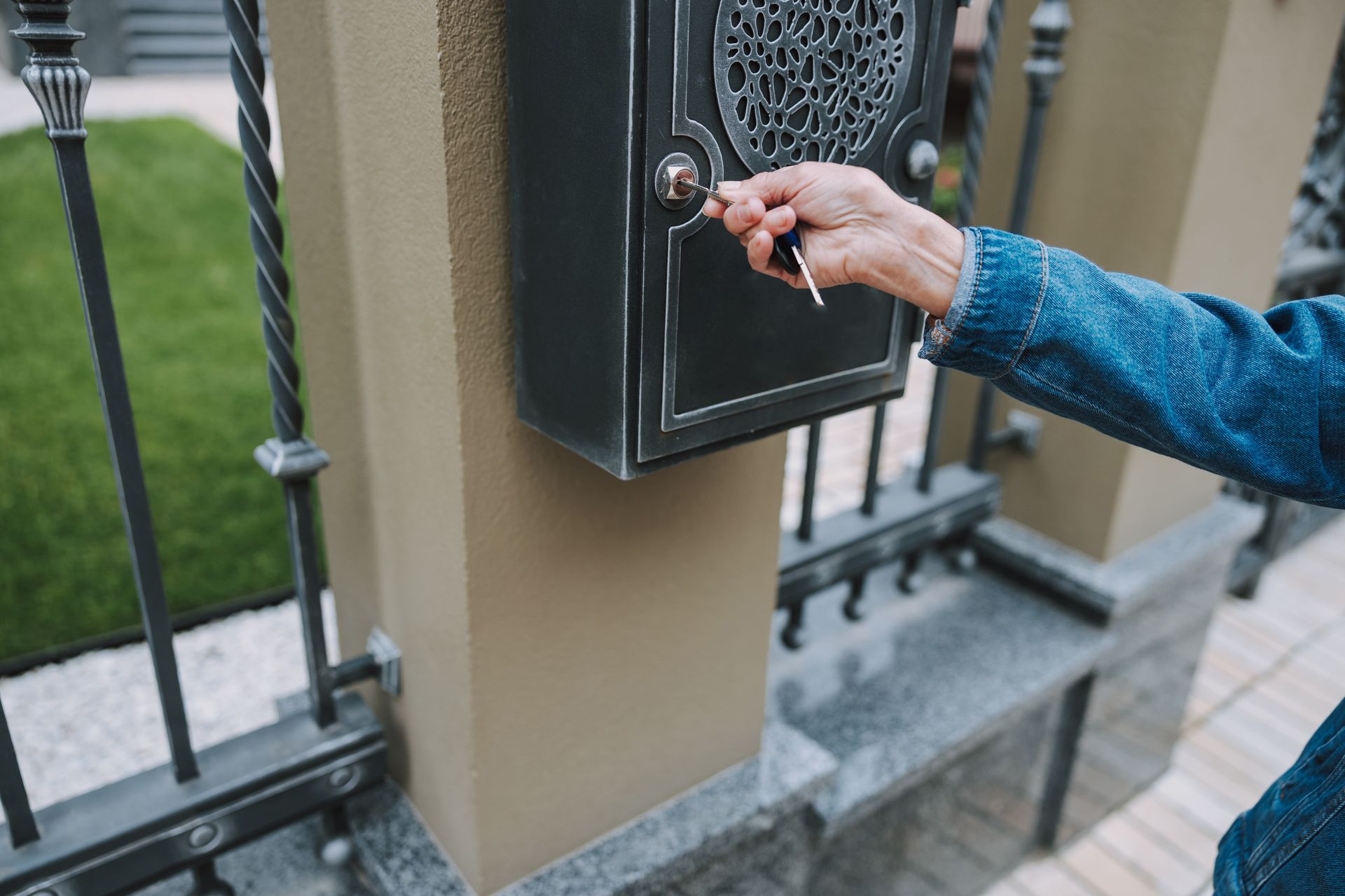 A person in a denim jacket uses a key to unlock a decorative black metal mailbox attached to a tan pillar and iron fence.