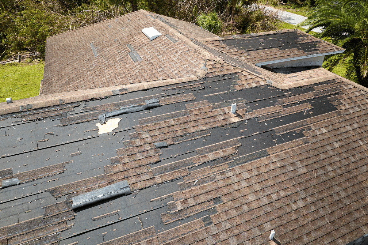 Damaged brown shingle roof with missing sections, revealing dark underlayment, in an outdoor setting.