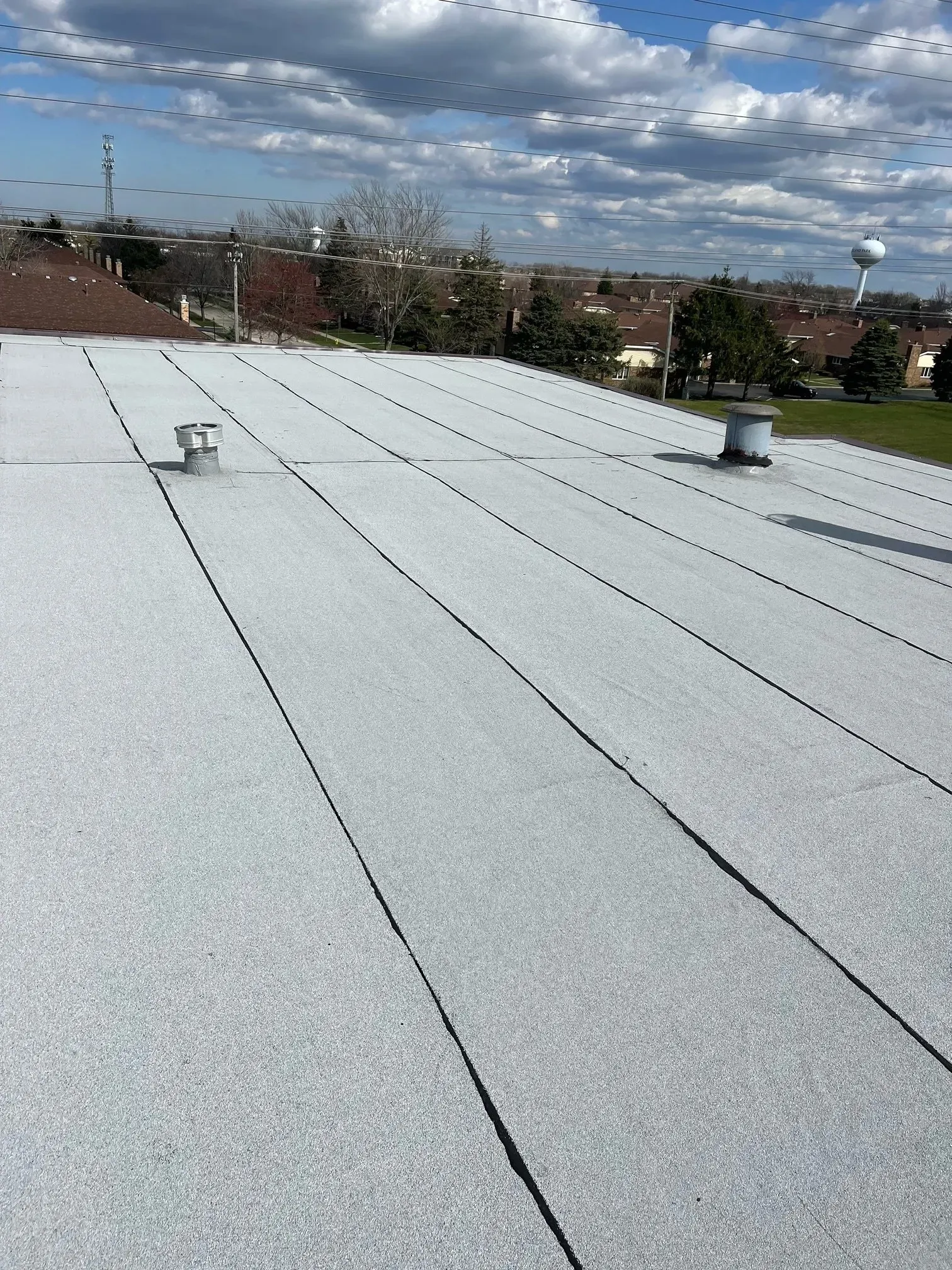 White commercial flat roof with black seams, vents, and surrounding buildings under a cloudy sky.