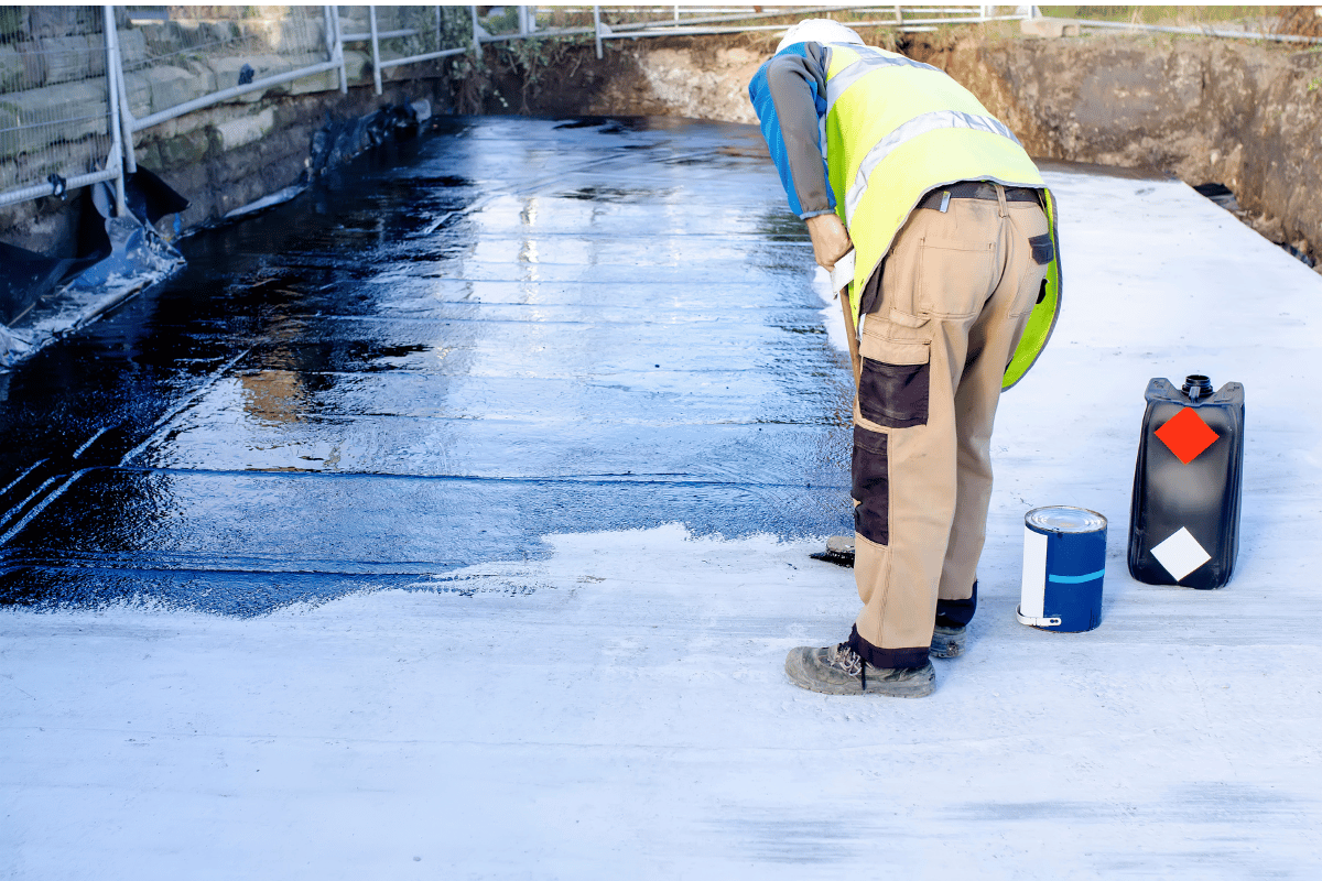 Person in workwear applying black sealant to a concrete surface.