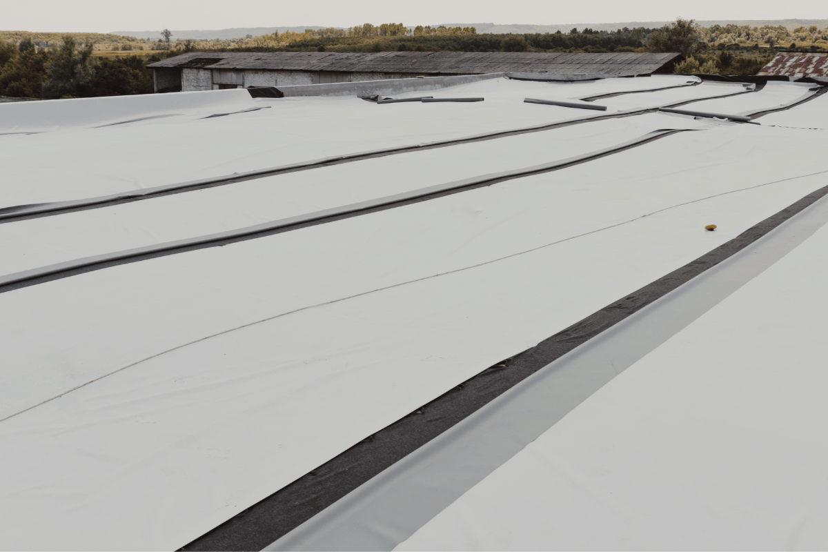Aerial view of a flat roof, covered in light-colored material, with dark seams, and a distant building.