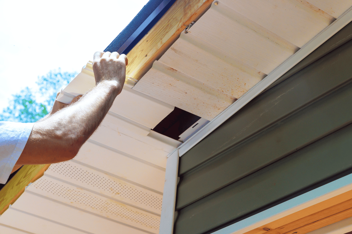 Person installing soffit vent on a house, with white siding and green corner.