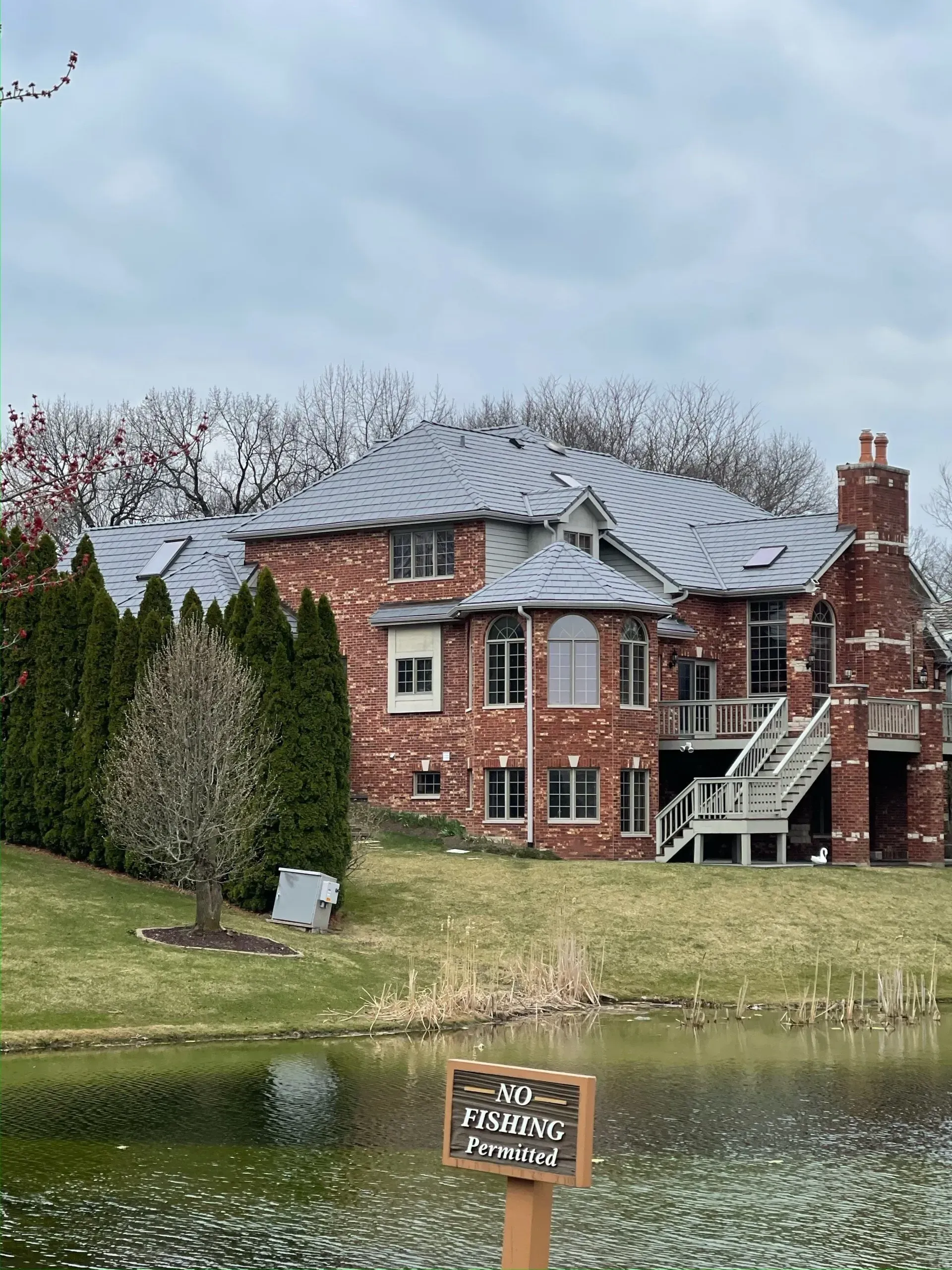 Large brick house with grey roof and deck overlooking a pond, 'No Fishing' sign in foreground.