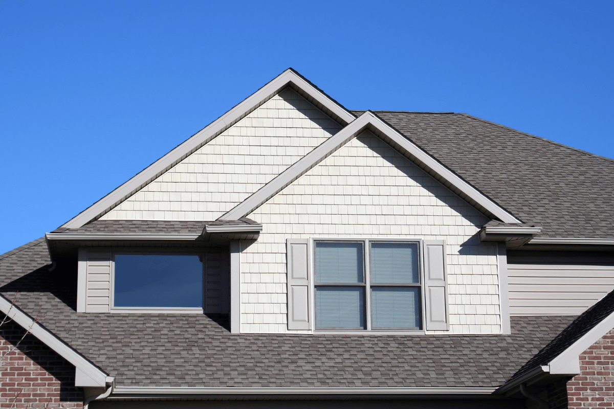 Close-up of a house with a brown roof, blue sky, and tan siding.