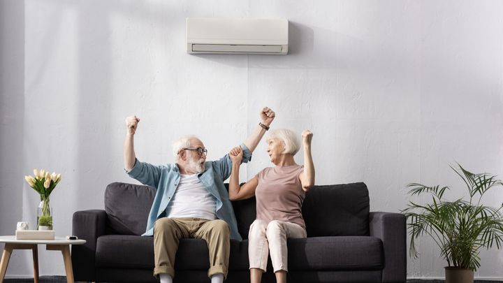 An elderly couple is sitting on a couch with their arms in the air.