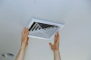 Hands installing a white air vent cover on a white ceiling.