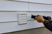 Person caulking around a white dryer vent on a white siding exterior wall.
