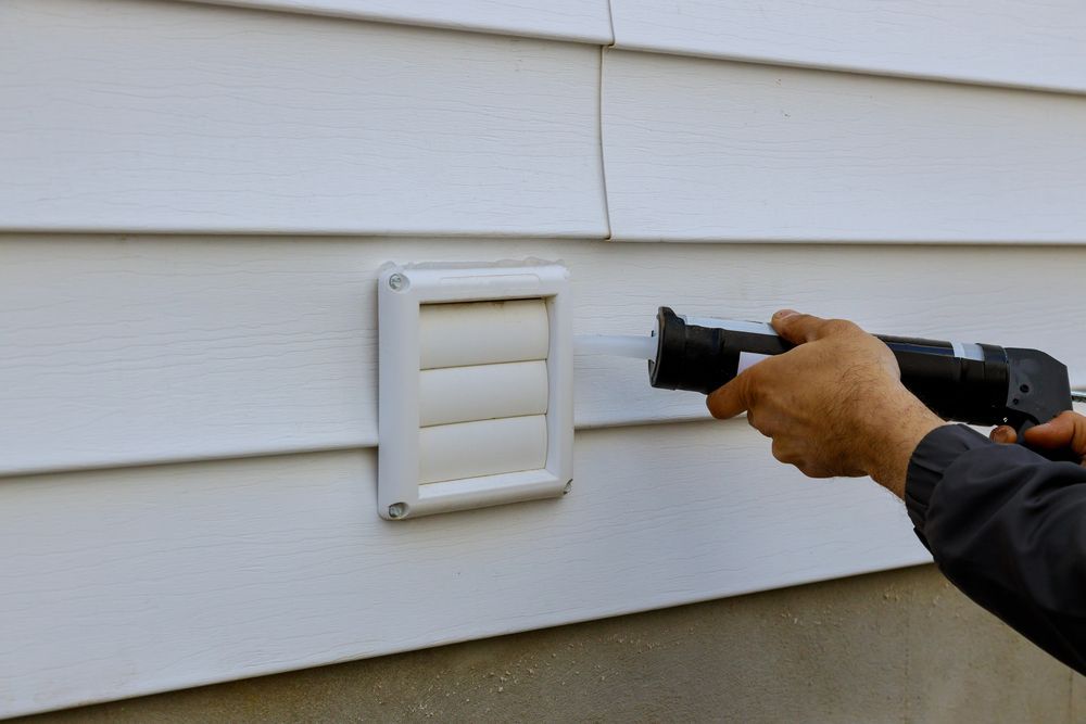 Person caulking around a white dryer vent on a white siding exterior wall.
