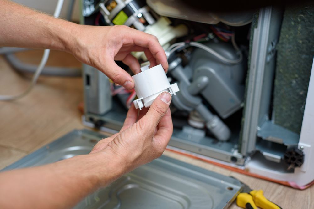 Person holding a white dishwasher part, working on the appliance with the front panel open.